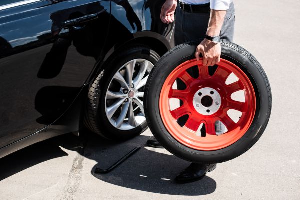 Człowiek niosący koło samochodowe, obok stoi auto