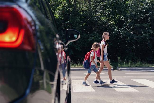 Dwójka dzieci przechodząca przez przejście dla pieszych. Przed pasami znajduje się samochód przepuszczający pieszych
