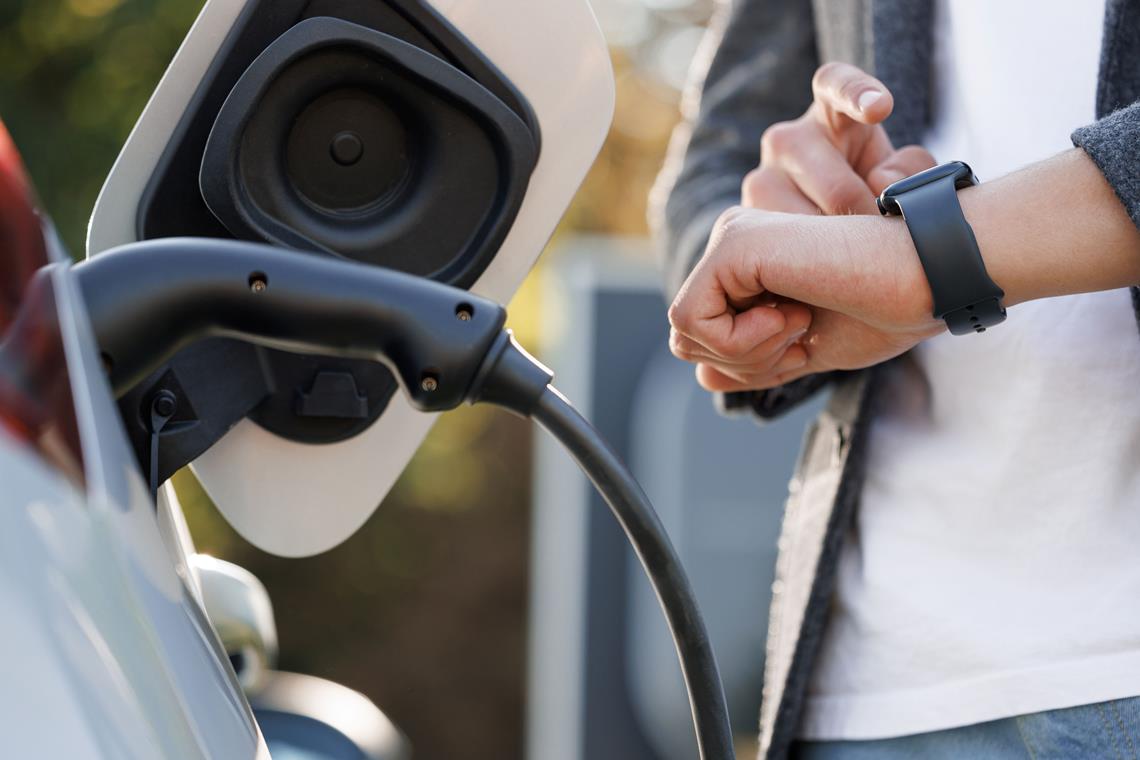 mężczyzna zerkający na zegarek stojący przy samochodzie elektrycznym, który podpięty jest do stacji ładowania